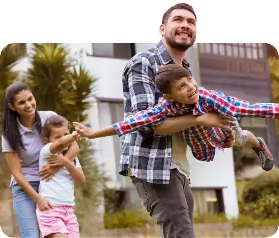 A happy family enjoying time outside their home with a strong, reliable roof overhead. The father playfully lifts his son like an airplane, while the mother and daughter smile nearby, showcasing the safety and comfort a well-built roof provides.